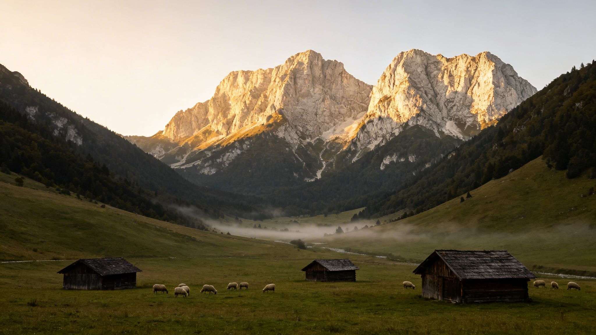 Majestic mountains bathed in golden light at sunrise, overlooking a misty valley with grazing sheep and wooden cabins.