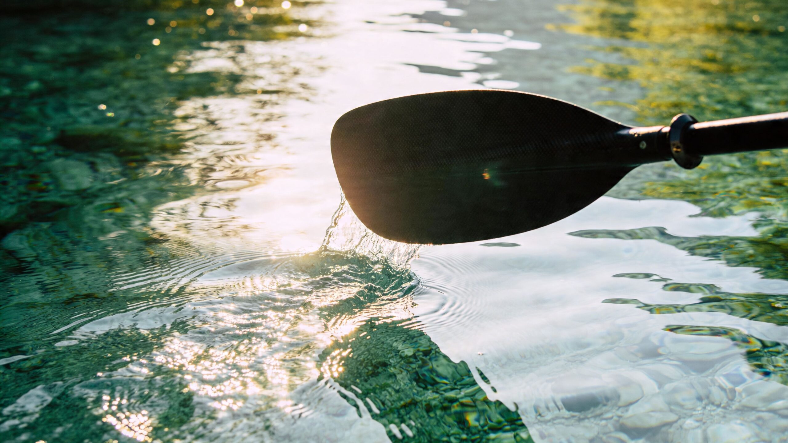 A close-up view of a black paddle blade slicing through clear turquoise water on a sunny day.
