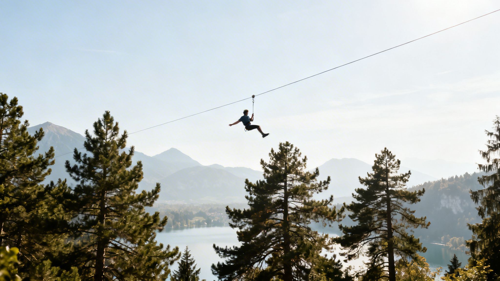 A person ziplining over a scenic lake, surrounded by mountains and tall pine trees.