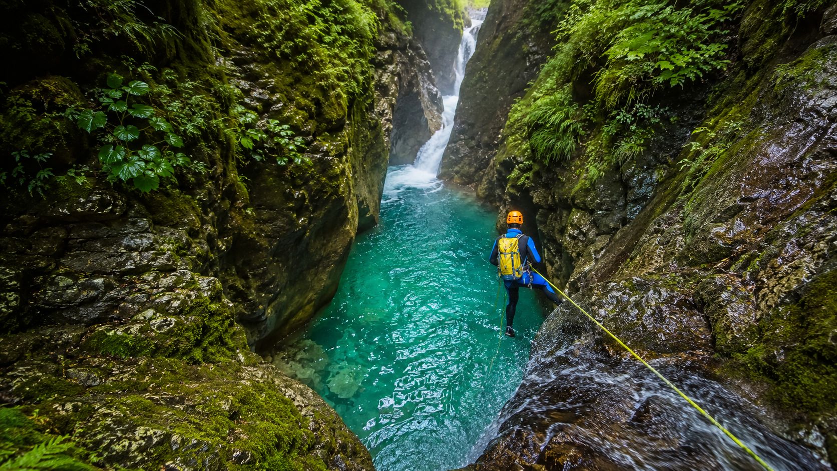 A person in a wetsuit and helmet rappelling down a waterfall in a lush, rocky canyon.