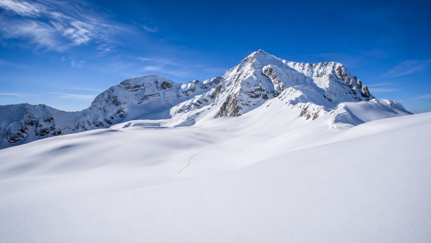 A vast, sunlit snowy landscape with jagged mountain peaks under a clear blue sky.