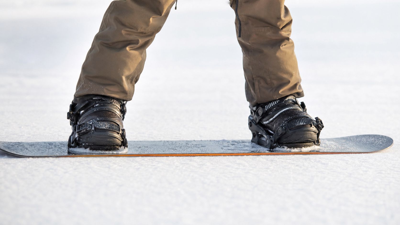 A close up view of a snowboarder wearing tan pants and black bindings on snow-covered ground.