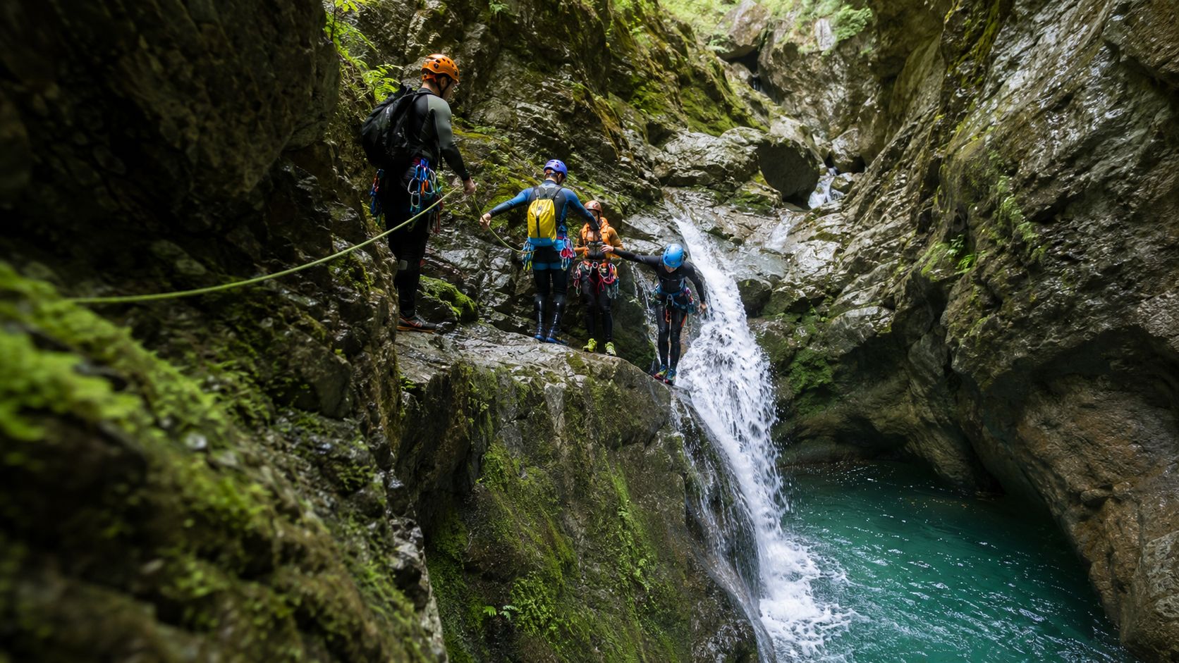 A group of people wearing helmets and wetsuits participating in canyoning activities near a mountain waterfall