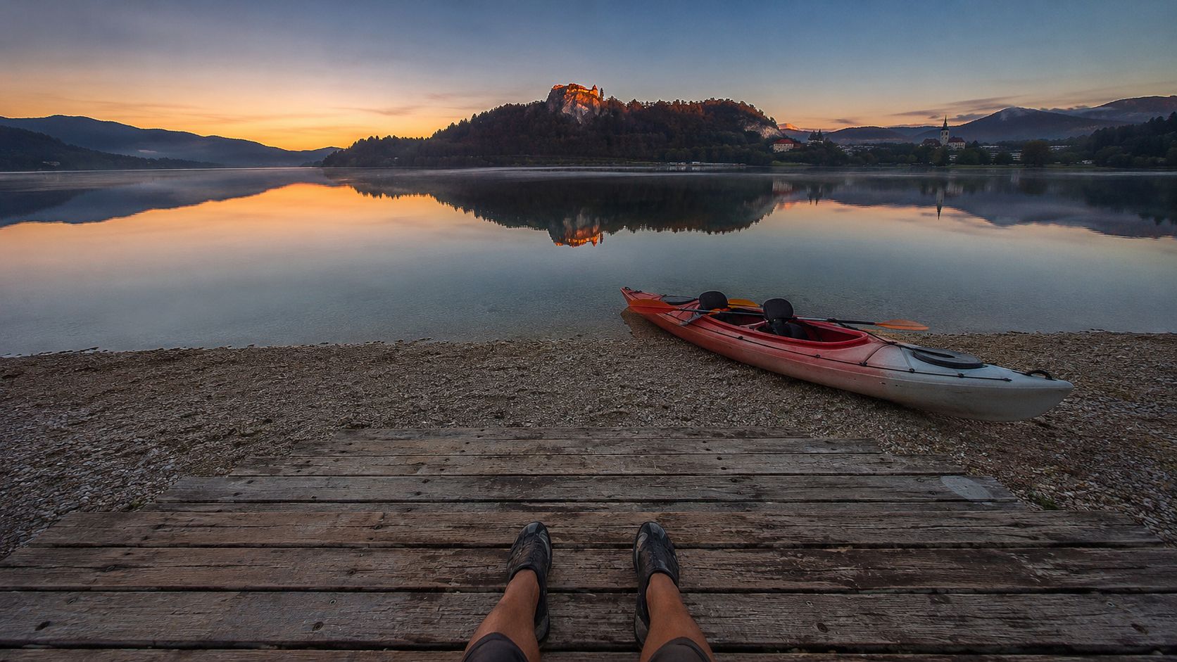 A person sitting on a wooden dock overlooking Lake Bled at sunrise with a red kayak nearby.